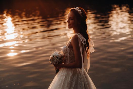 An elegant bride in a white dress and gloves stands by the river in the Park with a bouquet, enjoying nature at sunset.A model in a wedding dress and gloves in a nature Park.Belarus.