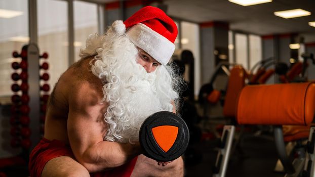 Santa claus in the gym. Muscular man with a naked torso doing exercises with dumbbells