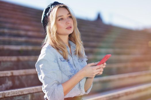 Blonde woman using her smartphone sitting on some city steps, wearing a denim shirt and black beret.