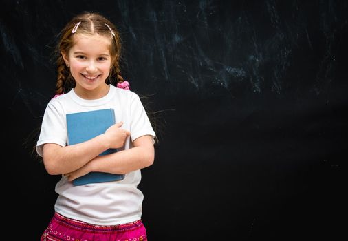 cute little girl smiling on black school board background with a blue book in hand