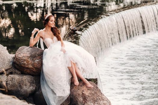 An elegant bride in a white dress, gloves and bare feet is sitting near a waterfall in the Park enjoying nature.A model in a wedding dress and gloves at a nature Park.Belarus.