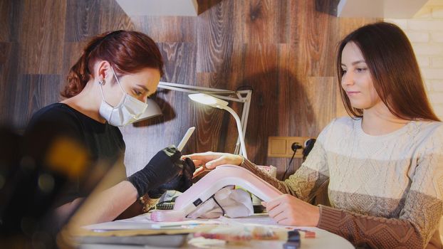 Manicurist in medical mask doing manicure for attractive woman in beauty salon, horizontal