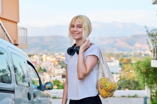 Portrait of female teenager with oranges in eco-friendly mesh bag. Beautiful smiling blonde teen girl with headphones looking at camera, mountains sunset city. Adolescence, trends, healthy food