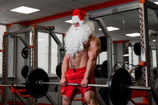 Santa claus in the gym. Muscular man with a naked torso doing exercises with dumbbells