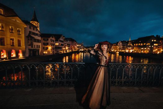 A stylish bride in a black wedding dress and red hat poses at night in the old city of Zurich. Portrait of a model girl after sunset.Photo Shoot In Switzerland