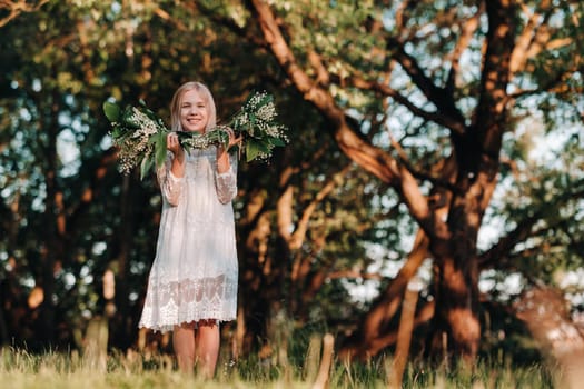 A beautiful nine-year-old blonde girl with long hair in a long white dress, holding a bouquet of lilies of the valley flowers, walking in nature in the Park.Summer, sunset