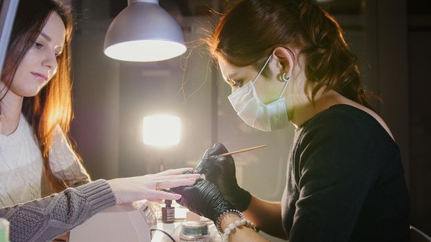 Nail master in medical mask doing professional manicure for white caucasian model, backlight, close up