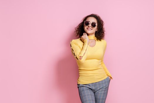 Enjoying herself. photo of a beautiful young curly caucasian girl in a yellow outfit , who is laughing and posing with her right hand over her head on pink background . Looking at camera