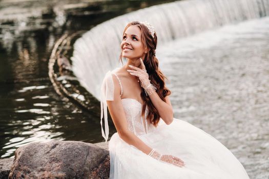 An elegant bride in a white dress, gloves and bare feet is sitting near a waterfall in the Park enjoying nature.A model in a wedding dress and gloves at a nature Park.Belarus.