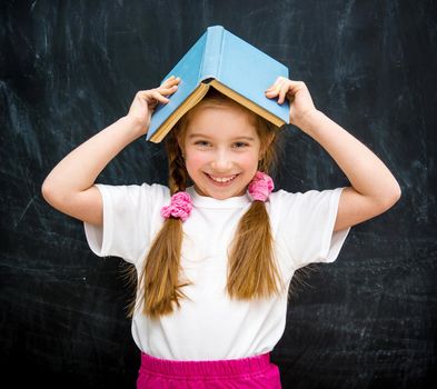cute little girl with a blue book on her head smiling on the background of a school blackboard