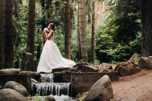 An elegant bride in a white dress and gloves holding a bouquet stands by a stream in the forest, enjoying nature.A model in a wedding dress and gloves in a nature Park.Belarus.