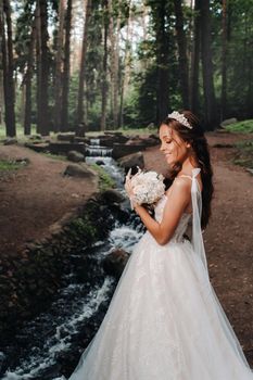 An elegant bride in a white dress and gloves holding a bouquet stands by a stream in the forest, enjoying nature.A model in a wedding dress and gloves in a nature Park.Belarus.