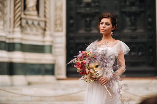 the girl-bride is with beautiful flower pattern as a mask in Florence, stylish bride in a wedding dress standing with a mask in the Old town of Florence. Model girl in Florence.