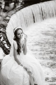 An elegant bride in a white dress, gloves and bare feet is sitting near a waterfall in the Park enjoying nature.A model in a wedding dress and gloves at a nature Park.Belarus.black and white photo