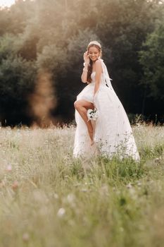 On the wedding day, an Elegant bride in a white long dress and gloves with a bouquet in her hands stands in a clearing enjoying nature. Belarus