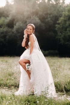 On the wedding day, an Elegant bride in a white long dress and gloves with a bouquet in her hands stands in a clearing enjoying nature. Belarus