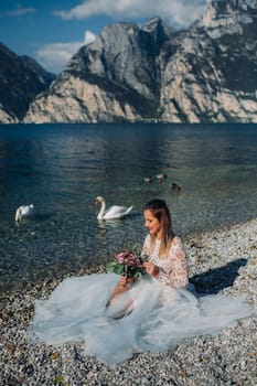 a girl in a smart white dress is sitting on the embankment of lake Garda.A woman is photographed against the background of a mountain and lake in Italy.Torbole.