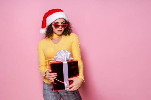 Young attractive curly girl in Santa's hat and red sunglasses holds red giftbox while stands on pink background. New Year concept