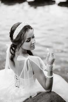 An elegant bride in a white dress and gloves is sitting by the lake in the Park, enjoying nature.A model in a wedding dress and gloves in a nature Park.Belarus.black and white photo.