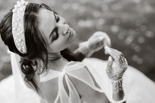 An elegant bride in a white dress and gloves is sitting by the lake in the Park, enjoying nature.A model in a wedding dress and gloves in a nature Park.Belarus.black and white photo.