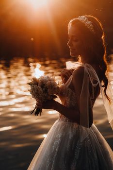 An elegant bride in a white dress and gloves stands by the river in the Park with a bouquet, enjoying nature at sunset.A model in a wedding dress and gloves in a nature Park.Belarus.