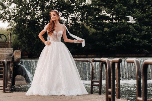 An elegant bride in a white dress and gloves holding a bouquet stands by a stream in the forest, enjoying nature.A model in a wedding dress and gloves in a nature Park.Belarus.