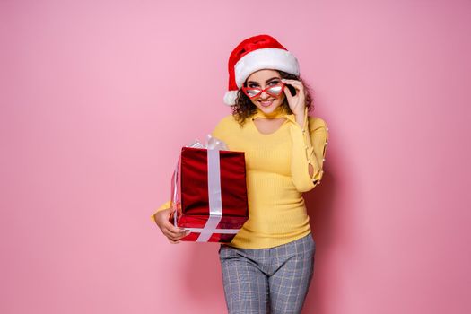 Young smiling happy cheerful curly girl in Santa's hat and red glasses holds red giftbox while stands on pink background. New Year concept