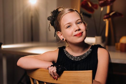 Stylish little girl in a black dress sits on a chair in an evening interior.