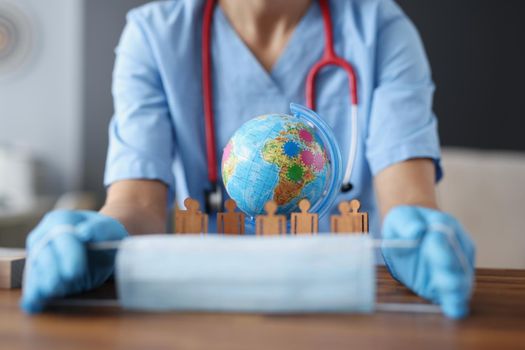 Close-up of female doctor hands holding sterile mask. Globe and wooden person symbols on desk. Worldwide pandemic, covid-19 and coronavirus outbreak concept