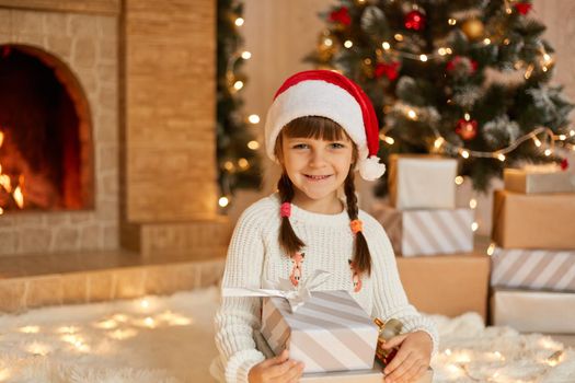 Cute female child in red hat on floor at home. Christmas celebration with presents, little girl posing with gift boxes, looks smiling at camera, dresses white jumper, sits near fireplace and xmas tree