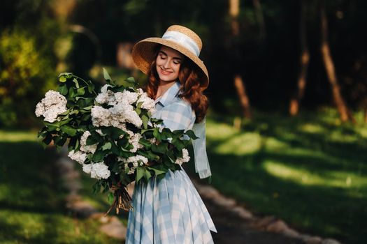 Portrait of a beautiful girl with long hair, a straw hat and a long summer dress with lilac flowers in the garden.