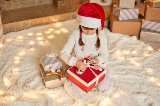 Little Caucasian female child in santa hat and white jumper sitting on floor on soft carpet and packing Christmas gifts, looking concentrated on presents boxes, celebrating New Year.