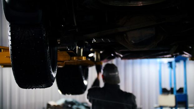 Car service - mechanic unscrewing automobile parts while working under a lifted car, backlight