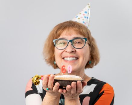 A portrait of a smiling elderly woman in a festive cap holding a cake with candles in the form of the number 60. Anniversary.