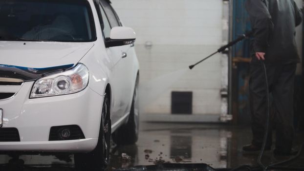 Mechanic in car service is washing automobile in the suds by water hoses, telephoto