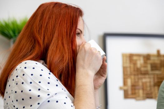 portrait of red-haired woman enjoys drinking coffee from a white cup in cafe. side view close up