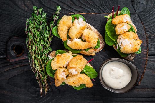 Toasts with Crispy Fried Shrimps Prawns and green salad on a wooden board. Black wooden background. Top view.