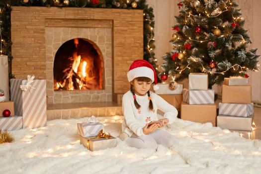 Charming girl communicates with relatives on phone via video call and thanks them for gifts, wearing white pullover and red santa hus hat, sitting neat fireplace with concentrated look.