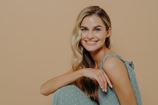 Fashion studio shot with smiling young woman on chair, charming pretty female in soft blue evening dress isolated over beige background with copy space, sitting sideways and looking at the camera