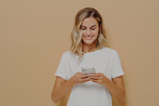 Young happy girl using smartphone, chatting with boyfriend in social media and smiling while standing against orange wall dressed in white t shirt. Modern technologies and networking concept