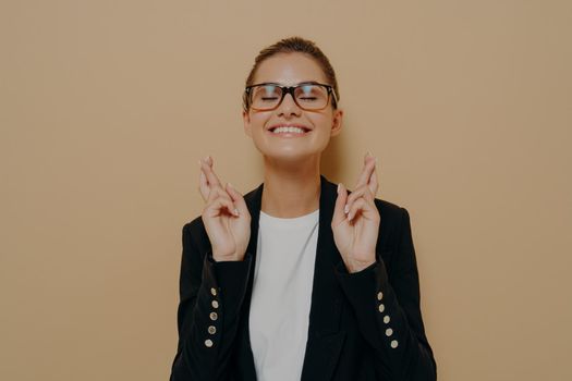 Young hopeful female student in eyeglasses crossing her fingers with hope and asking for good luck before exam, keeping eyes closed and smiling widely, dressed in casual wear. Body language concept