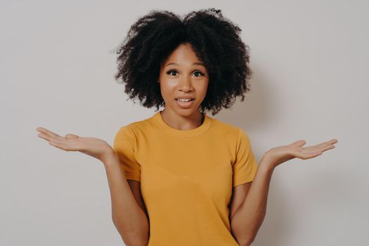 Confused doubtful funny black woman shrugging with shoulders, feeling baffled while looking at camera isolated on white blank studio background, clueless african female puzzled about new problem