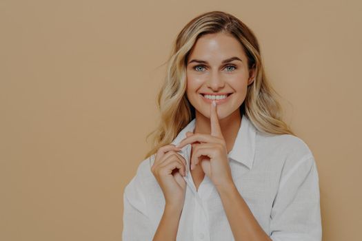 Keep it in secret. Young beautiful positive female with blond dyed hair touching lips with finger and smiling at camera, asking for silence while posing isolated over yellow wall in studio