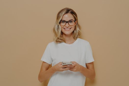 Young cute female student in spectacles using cell phone and looking at camera with pleasant smile, typing to friend or chatting in social media. Modern technologies and communication concept