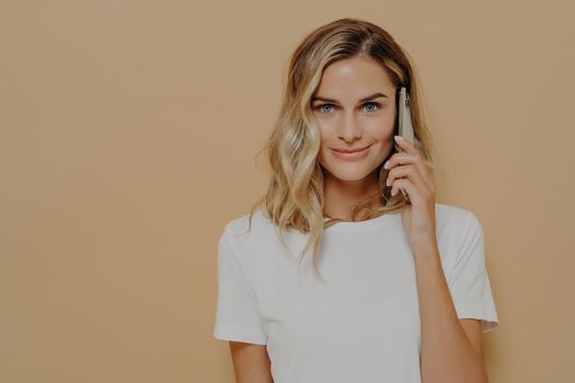Confident female with blonde dyed hair holding smartphone and calling to friend, looking at camera with smirk while posing in studio, dressed in white t shirt. Modern technologies and communication