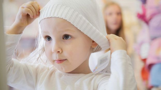 Child's dress store - little blonde baby girl with mother doing shopping and buying hat, telephoto