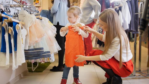 Cute little girl with mommy buying red dress in store of kids clothes , telephoto