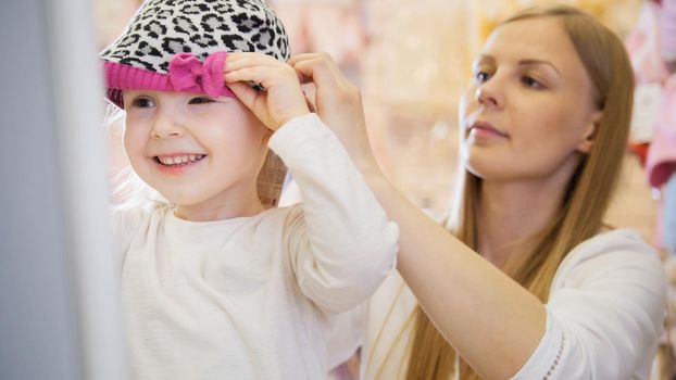 Kids dress store - little blonde baby girl with mother doing shopping and buying red hat, telephoto