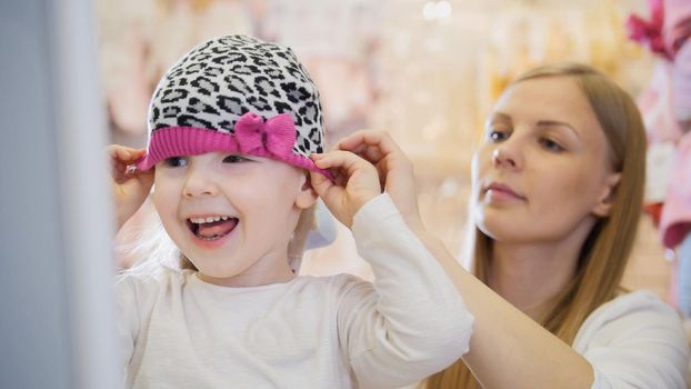 Little happy girl with mommy doing shopping and buying pink hat in kids dress store, telephoto