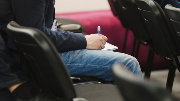 Student in auditorium writes in notebook with pen, close up, telephoto